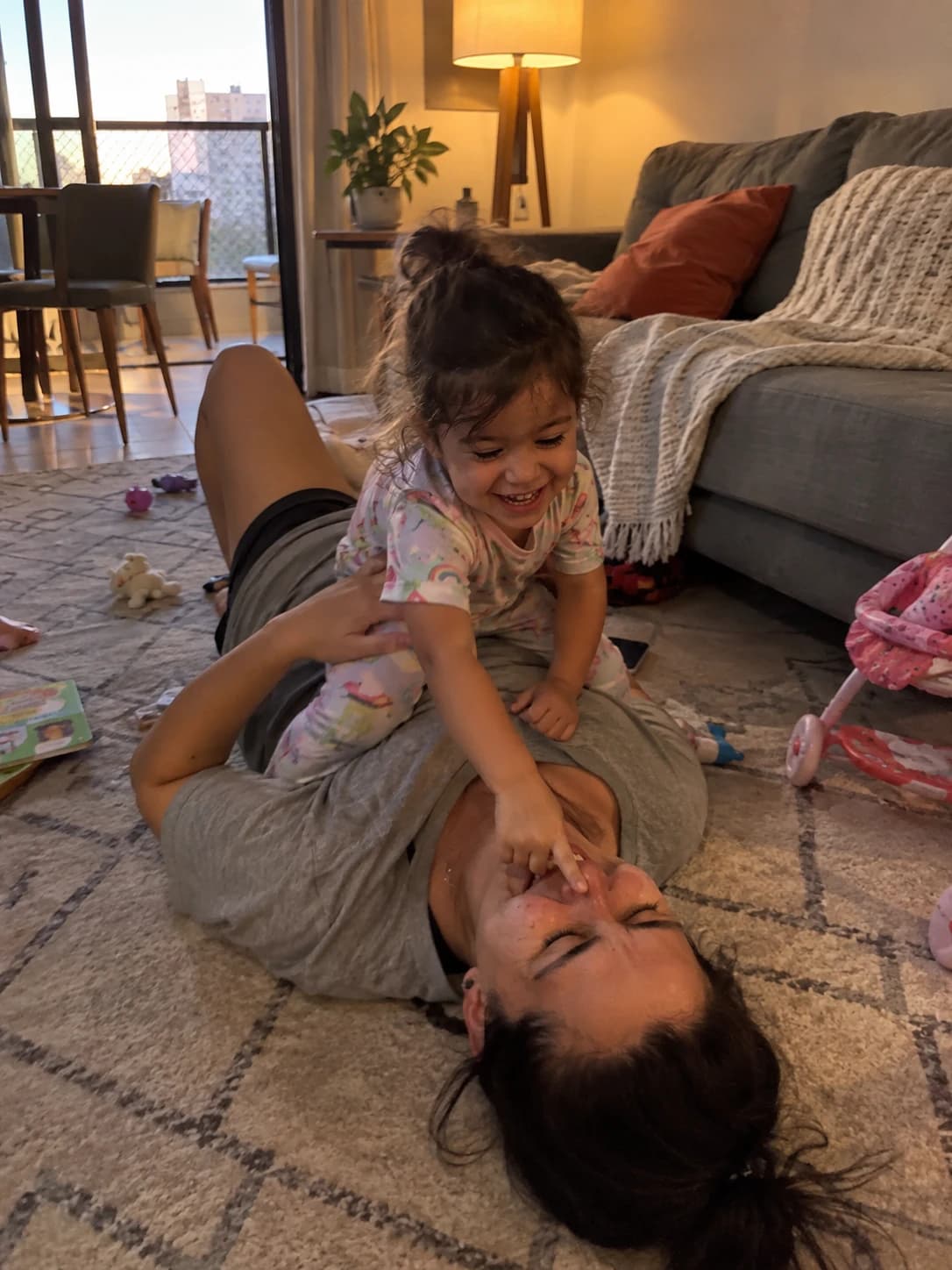 Mãe deitada no tapete da sala rindo, com a filha pequena sentada em cima apertando o nariz dela